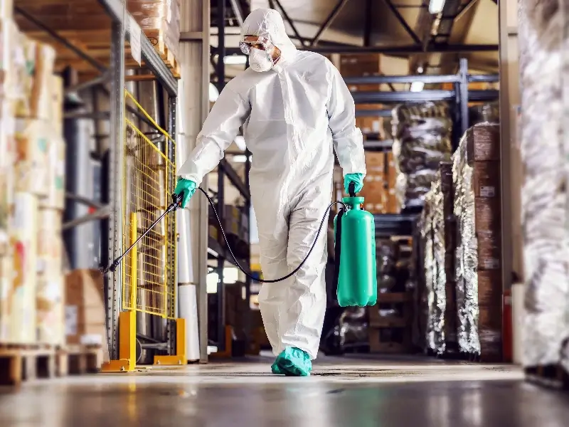 Person in protective suit spraying disinfectant in a warehouse aisle.