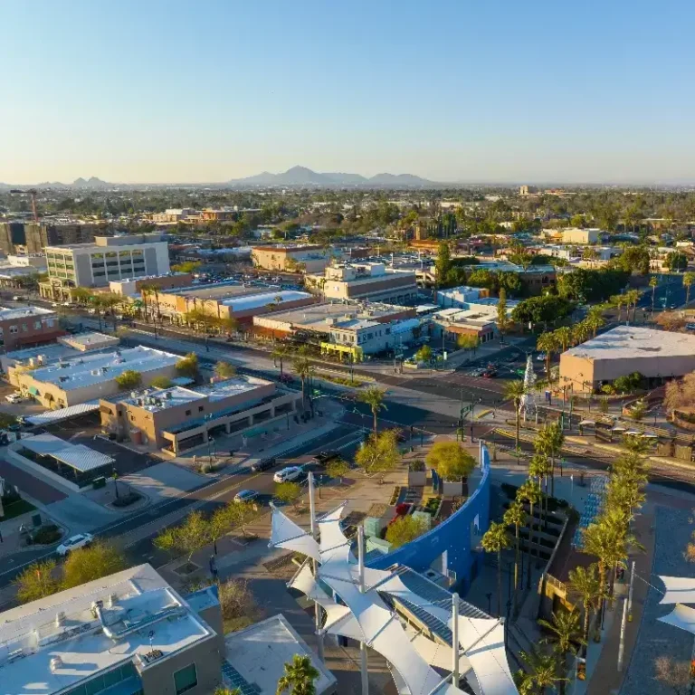 Aerial view of a cityscape with buildings, streets, palm trees, and distant mountains under a clear sky.