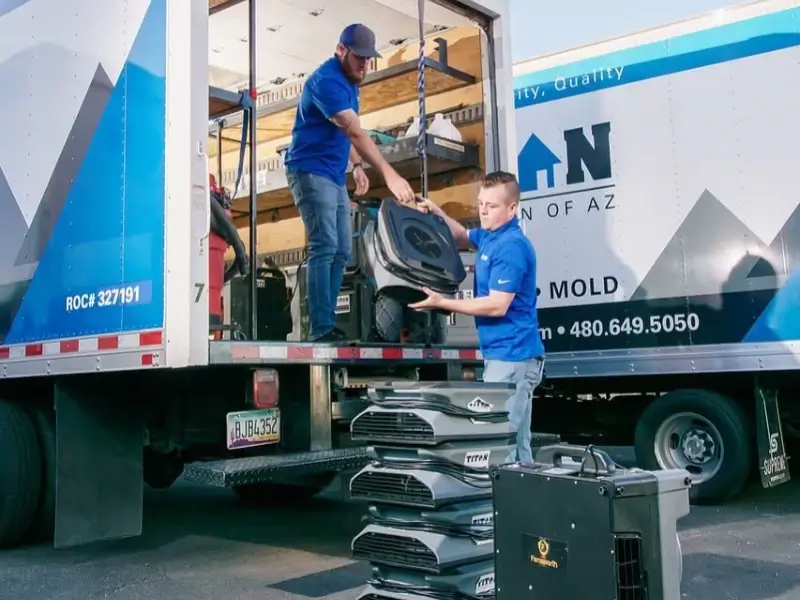 Two men in blue shirts unloading equipment from a truck with stacked air movers on the ground.
