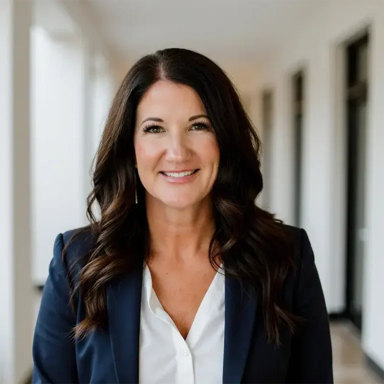 Smiling woman with long dark hair wearing a navy blazer and white blouse in a bright hallway.