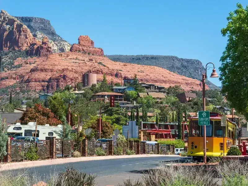 Yellow trolley on a road with red rock formations and houses in the background.