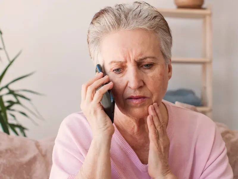 Older woman with short gray hair looks worried while talking on a phone and touching her cheek.