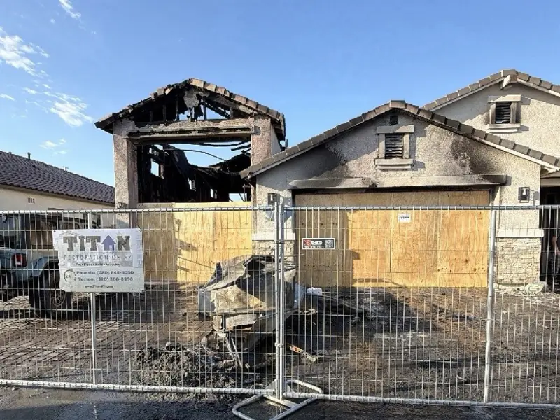House severely damaged by fire with boarded-up windows and a metal fence in front.