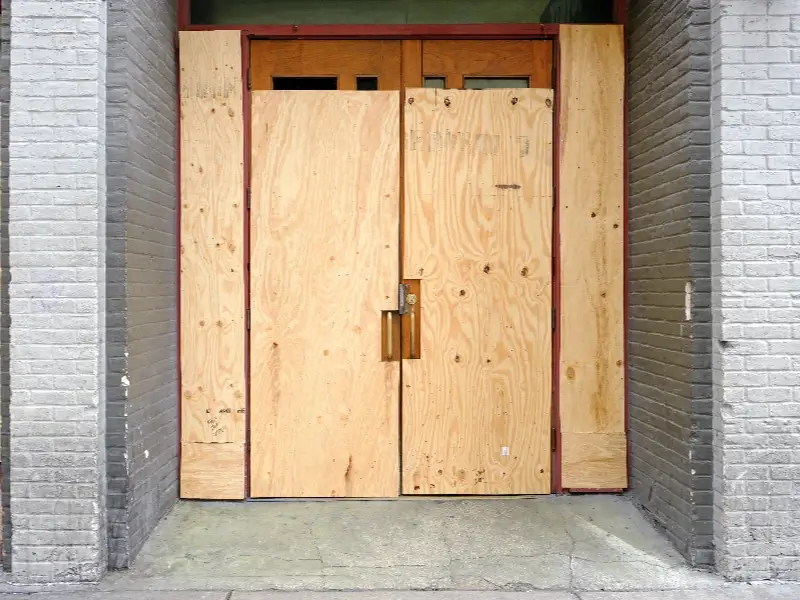 Double wooden doors boarded up with plywood within a gray brick building entrance.