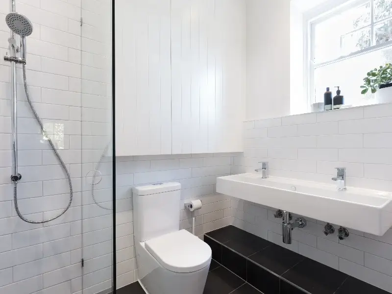 Modern bathroom with white toilet, double sink, glass shower, and black tiled floor steps.