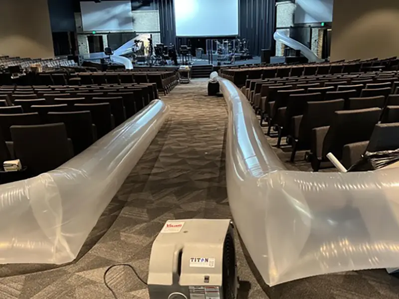 Two large inflatable tubes connected to a blower in an empty auditorium with rows of seats.