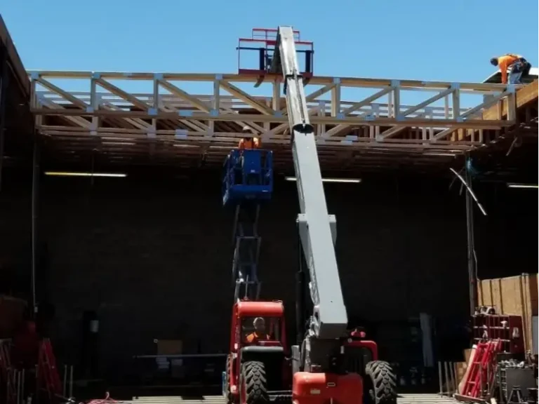 Construction workers on a wooden roof frame using a red boom lift inside a building.