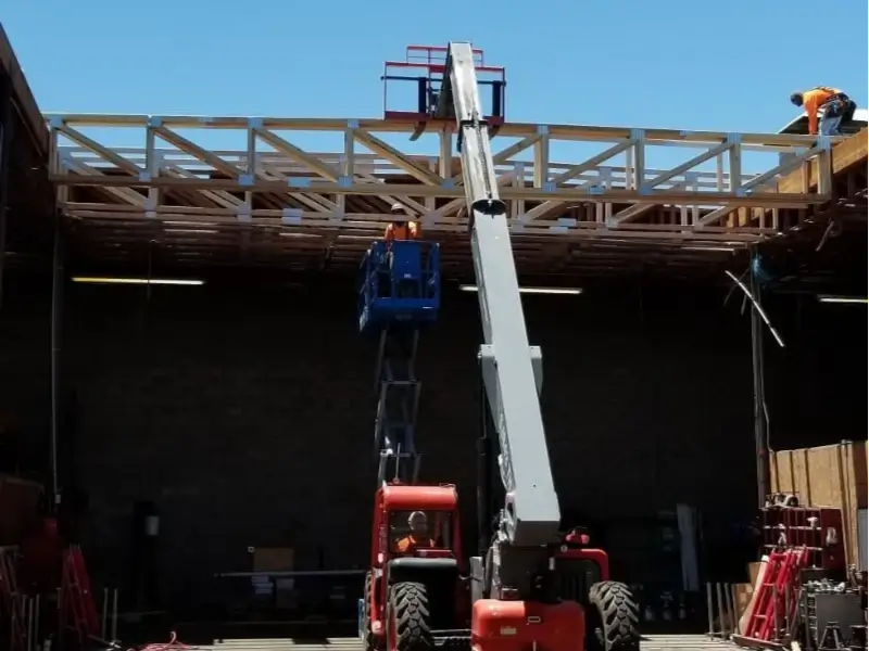 Construction workers on a wooden roof frame using a red boom lift inside a building.