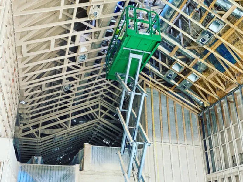 Green scissor lift raised inside a building under construction with exposed roof trusses and ductwork.