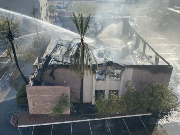 Firefighters spray water on a partially burned building with smoke rising and palm trees nearby.