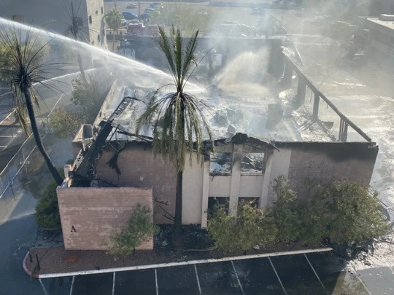 Firefighters spray water on a partially burned building with smoke rising and palm trees nearby.