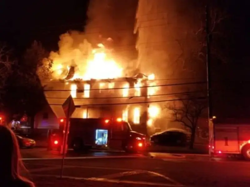 A house engulfed in flames at night with fire trucks and emergency lights nearby.