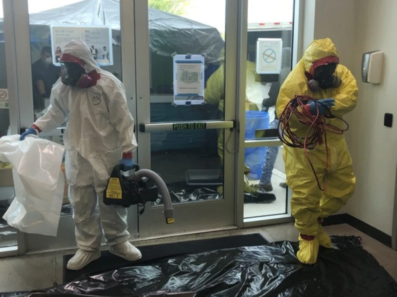 Two people in hazmat suits, one white and one yellow, preparing equipment inside a building.