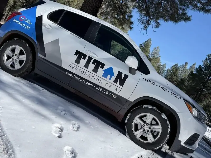 White and blue Titan Restoration of AZ service vehicle parked on snow-covered ground near trees.