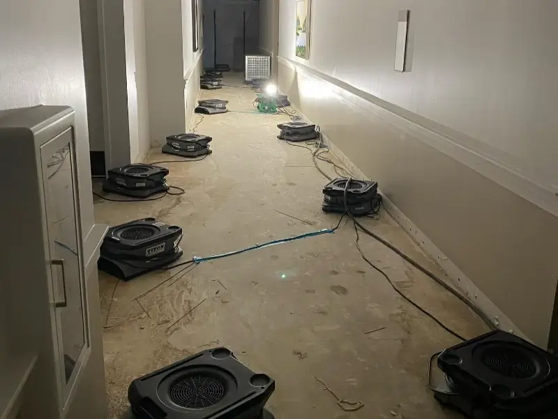 Multiple air movers arranged along a hallway with a bare floor and exposed wires.