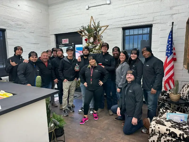 Group of people in black jackets and hats posing indoors near a decorated Christmas tree and American flag.