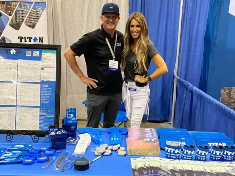 Two people stand behind a table with blue promotional items and a display board at a booth.