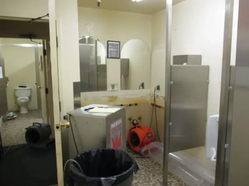 Restroom under maintenance with drying equipment, trash can, and open toilet stall visible.