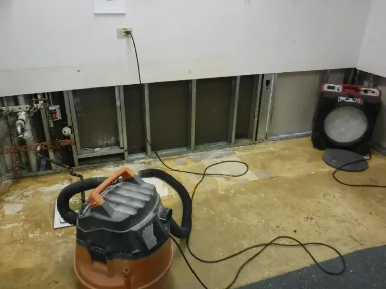 Room with exposed wall studs, a wet/dry vacuum, and a drying fan on a bare floor.