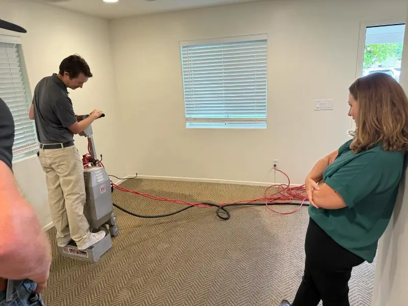 A man operates a floor cleaning machine while a woman watches leaning against a wall in a carpeted room.