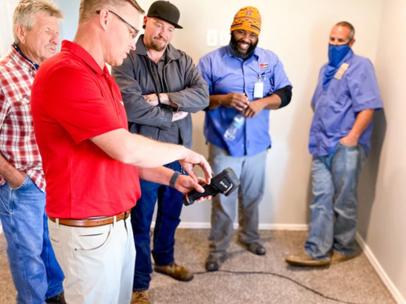 A man in a red shirt demonstrates a device to four men watching in a room with carpeted floor.