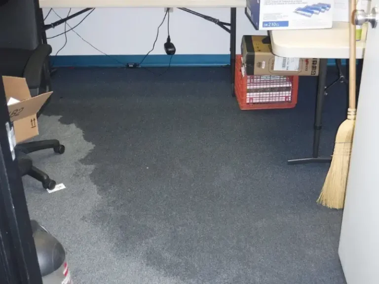 Office floor with a large wet stain near a chair and a broom leaning against a table.